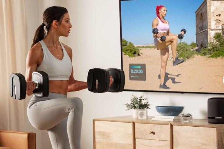 Woman uses NordicTrack adjustable dumbbells in front of iFit fitness app on TV