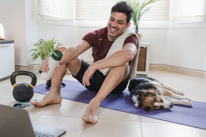 Man sits on exercise mat looking at open laptop, a kettlebell is next to him