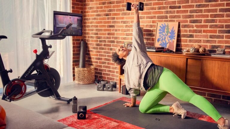 Woman performing dumbbell exercise on yoga mat. A Peloton Bike+ is next to her with the in-built screen turned her way and showing a fitness class